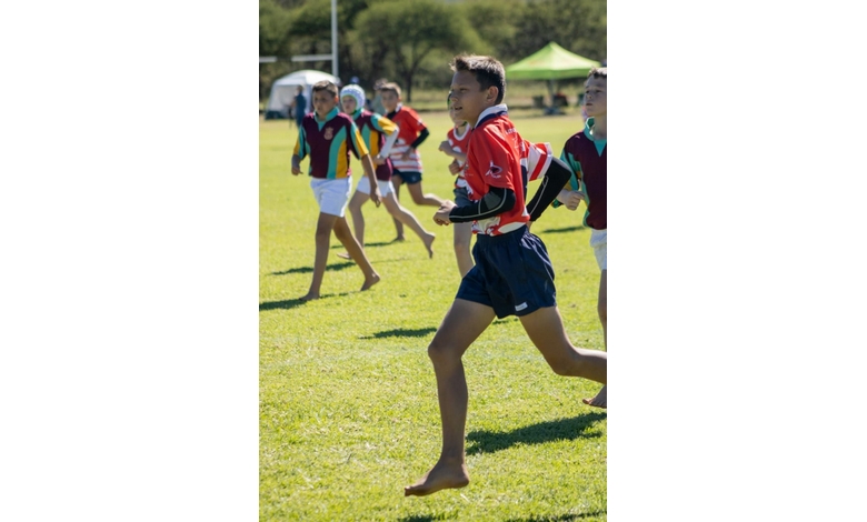 A group of young boys are playing rugby outside on a sunny day. One boy in a red jersey is running barefoot with determination, while others follow.