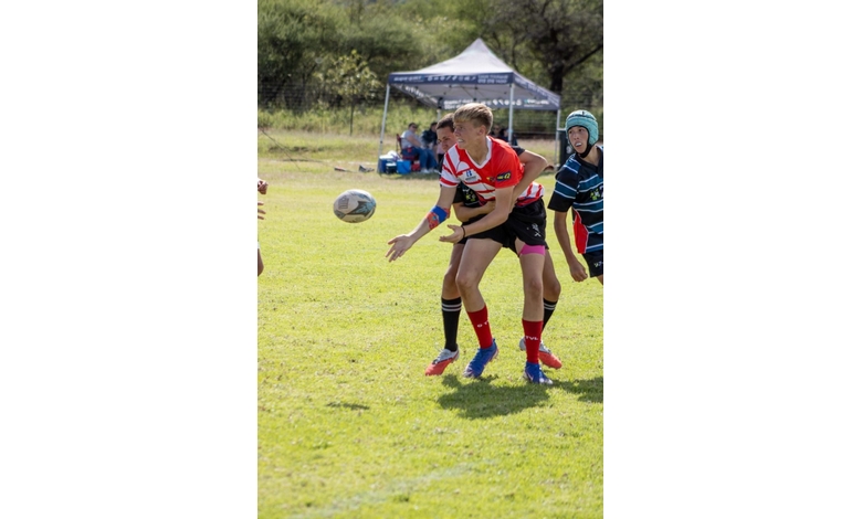 A rugby player in a red and white jersey passes the ball mid-game, concentrating intensely. Another player in a black and blue jersey follows closely behind, with a tent visible in the background. The scene conveys action and focus.