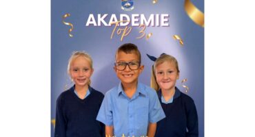 Three smiling children in school uniforms stand in front of a blue backdrop with "AKADEMIE Top 3" written above. Gold confetti decorates the image.