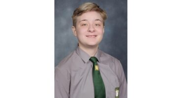 Smiling person with short light brown hair wearing a gray school uniform shirt and green tie against a gray-blue backdrop, exuding a cheerful vibe.