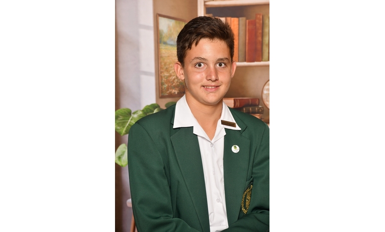 A young person in a green blazer and white shirt smiles warmly, standing in front of a bookshelf filled with books. The setting suggests a school photo.
