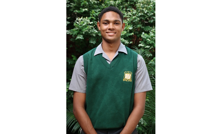 Young man in a green school uniform vest smiles confidently, standing before lush greenery, conveying a sense of calm and readiness.
