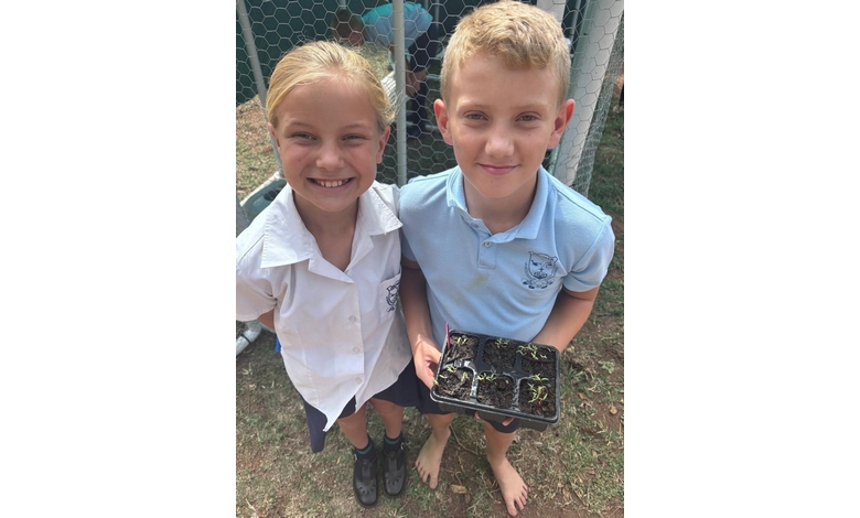 Two children in school uniforms stand outside, one holding a tray with small seedlings. They are smiling and standing on grass near a wire fence.
