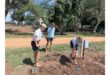 Three children in school uniforms work barefoot on a fenced-off patch of soil outdoors, with shovels and gardening tools, under sunny weather.