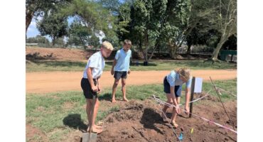Three children in school uniforms work barefoot on a fenced-off patch of soil outdoors, with shovels and gardening tools, under sunny weather.