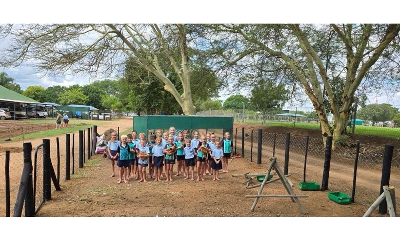 A group of children in blue uniforms stand in a fenced dirt area holding chickens. Trees and farm buildings are visible in the background. The scene is cheerful and lively.