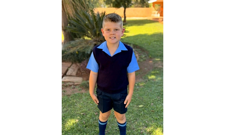 Young boy in a blue school uniform stands on a lawn, smiling confidently. Sunlight casts gentle shadows, creating a warm and cheerful atmosphere.