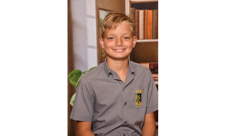 Smiling young boy in a gray button-up shirt with an emblem, seated indoors against a bookshelf backdrop, conveying a cheerful and studious vibe.