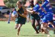 A young rugby player in a green and yellow uniform runs barefoot with the ball, pursued by opponents in blue. The scene is dynamic and intense.