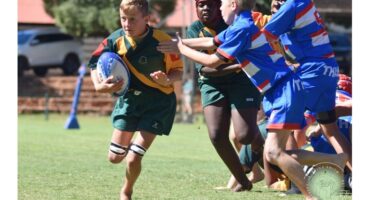 A young rugby player in a green and yellow uniform runs barefoot with the ball, pursued by opponents in blue. The scene is dynamic and intense.
