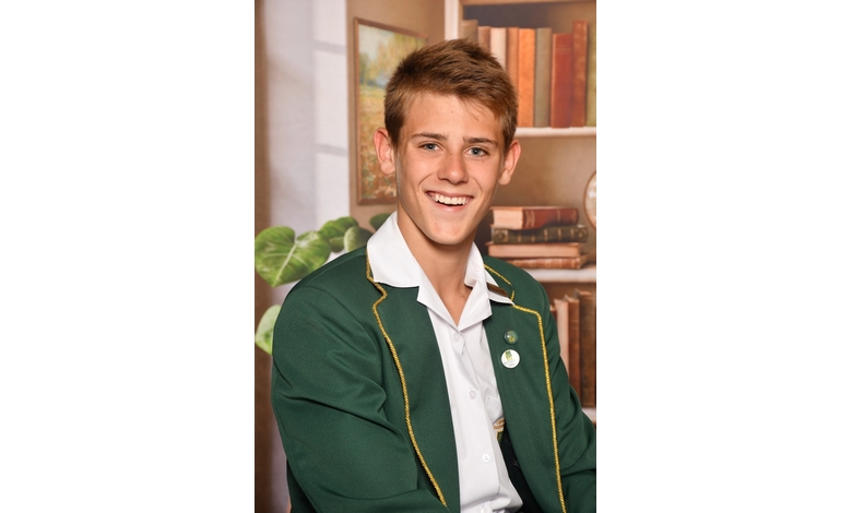 Smiling young man in a green blazer with gold trim, featuring badges. Background has a blurred bookshelf and a leafy plant, conveying a scholastic setting.