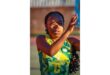 A netball player in a yellow and green uniform focuses intently while making a pass. Her braided hair swings with motion, set against a blurred outdoor court.