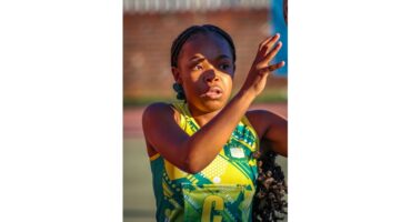 A netball player in a yellow and green uniform focuses intently while making a pass. Her braided hair swings with motion, set against a blurred outdoor court.