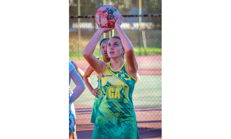 A netball player in a green and yellow uniform, marked "GA," aims to shoot a ball. Bright outdoor court setting, focus and intensity on her face.