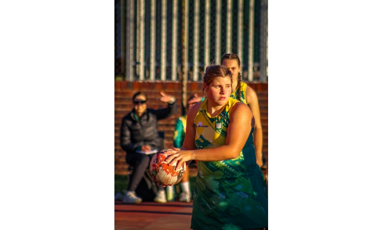 Young netball player wearing a green and yellow uniform holds a ball, focused on the game. Background features a blurred coach gesturing. Sunlight creates a warm tone.