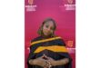 A woman in a graduation gown smiles softly in front of a red Stellenbosch University backdrop. Her expression conveys pride and accomplishment.