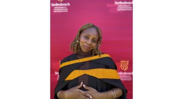 A woman in a graduation gown smiles softly in front of a red Stellenbosch University backdrop. Her expression conveys pride and accomplishment.
