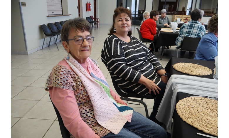 Two women sit at a table with woven placemats, smiling in a social gathering setting. The room includes other people seated at tables.