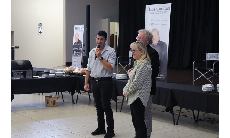 Three people stand in front of a stage with a man holding a microphone. Behind them are tables with food and a poster display, creating a formal event atmosphere.