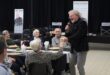 A smiling man with a microphone engages an elderly woman at a dining event, holding her hand. Other seniors look on with interest and joy.
