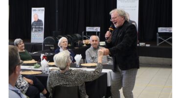 A smiling man with a microphone engages an elderly woman at a dining event, holding her hand. Other seniors look on with interest and joy.
