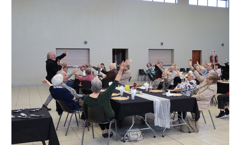 A group of elderly people sitting at decorated tables in a bright hall, all raising one hand in a cheerful gesture. A person stands at the front leading the activity.