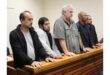 Five men stand in a courtroom, leaning on a wooden railing. They appear serious and attentive, each facing forward with a neutral expression.