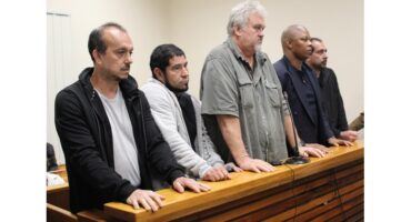 Five men stand in a courtroom, leaning on a wooden railing. They appear serious and attentive, each facing forward with a neutral expression.