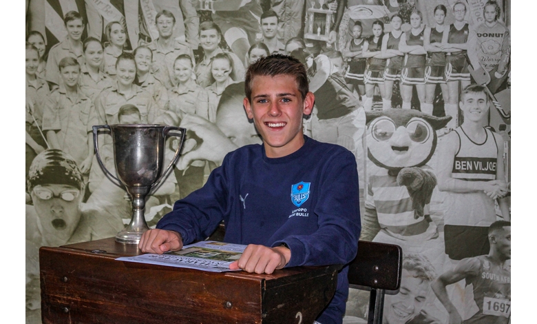 Young man smiling, sitting at a wooden desk with a sports trophy and brochures. Background features a collage of black-and-white sports photos.
