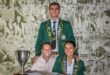 Three smiling people pose in front of a collage background. Two wear green blazers with badges, and one wears a light hoodie. A silver trophy is on the table.