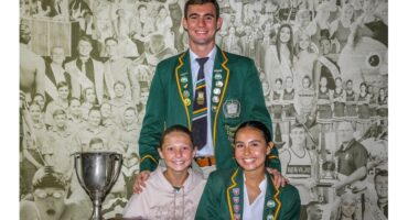 Three smiling people pose in front of a collage background. Two wear green blazers with badges, and one wears a light hoodie. A silver trophy is on the table.