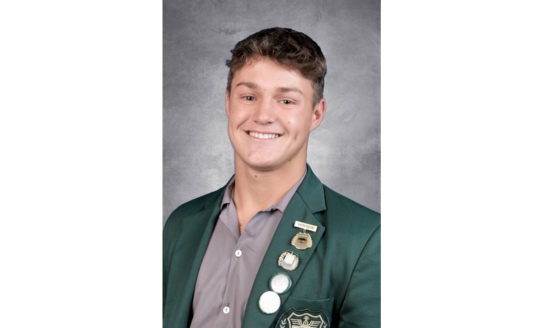 Young man smiling in a formal portrait, wearing a dark green blazer adorned with several pins. He stands against a light gray backdrop, conveying confidence.