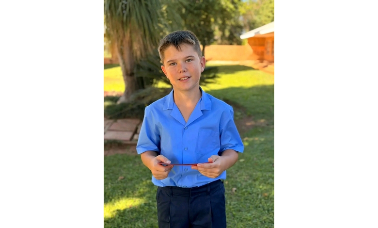 A young boy stands outside on a sunny day, wearing a blue shirt and dark pants. He holds a small object, smiling gently. Lush greenery surrounds him.