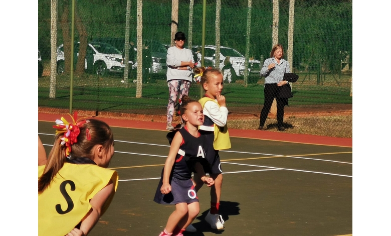 Young children energetically play netball on an outdoor court. They wear bright jerseys with letters. Bystanders watch near a green fence. Sunny weather.