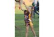A young girl in a brown and yellow sports uniform prepares to catch a ball during a game on a grassy field. She looks focused and determined.