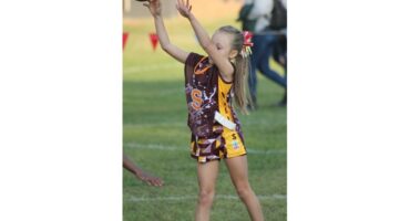 A young girl in a brown and yellow sports uniform prepares to catch a ball during a game on a grassy field. She looks focused and determined.