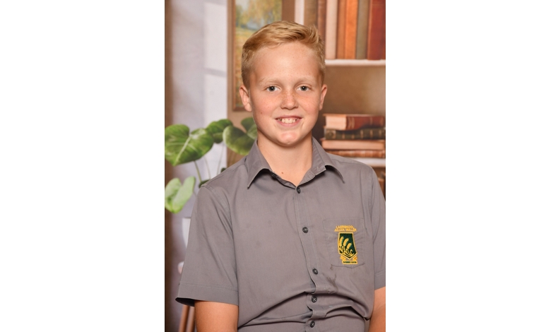 Smiling young person in a grey school uniform with an embroidered emblem, sitting against a backdrop of books and greenery. Warm, scholarly atmosphere.
