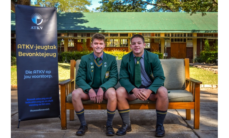 Two young men sitting on a bench, smiling, in front of a colorful banner displaying event details.