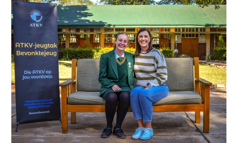 Two women relax on a bench, chatting, with a large sign visible in the background.