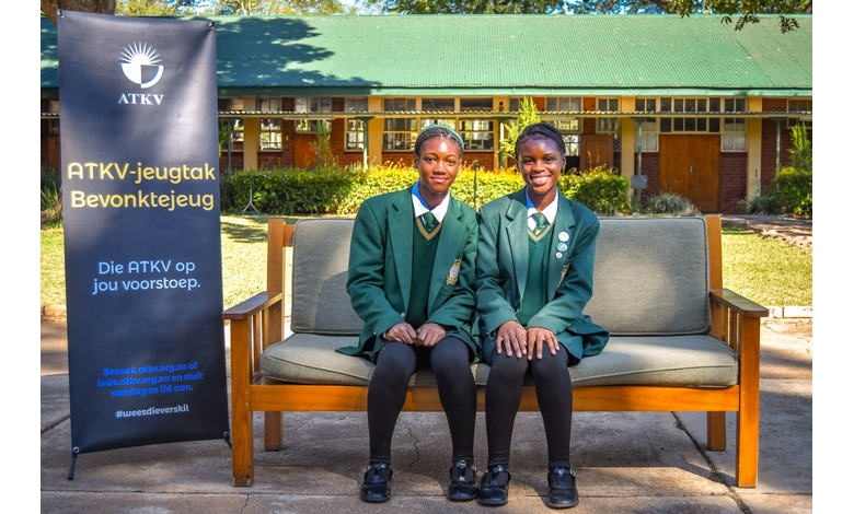 Two young girls sit on a bench, smiling in front of a colorful banner displaying a cheerful message.