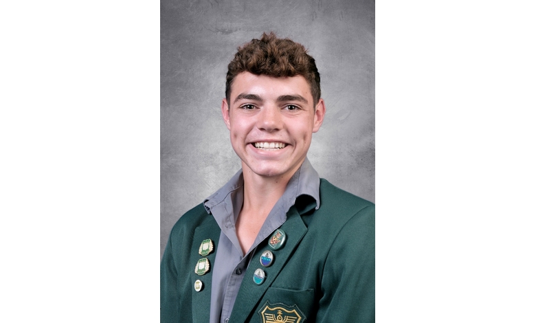 Smiling young person with short curly hair in a green blazer adorned with several colorful pins. The background is a simple gray texture.