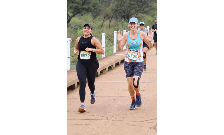 Two smiling runners on a scenic path during a race, one in black athletic wear and the other in colorful gear, surrounded by green trees.