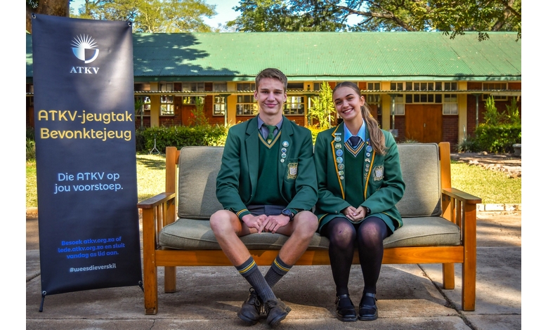A bench with two young individuals seated, with a large sign visible in the background, surrounded by greenery.