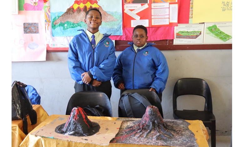 Two students in blue jackets proudly present volcano models on a table. Colorful educational posters adorn the wall, creating an academic atmosphere.