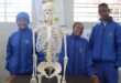 Three people in blue jackets stand smiling behind a skeleton model in a classroom. A chart is visible on the wall, suggesting an educational setting.