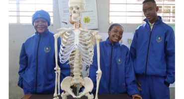 Three people in blue jackets stand smiling behind a skeleton model in a classroom. A chart is visible on the wall, suggesting an educational setting.