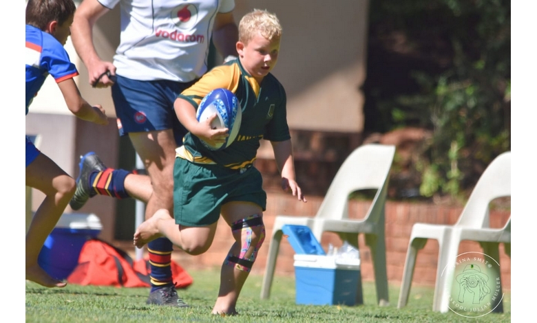 A young boy in a green and yellow rugby uniform runs with a rugby ball on a sunny day. He's focused, with teammates and chairs in the background.