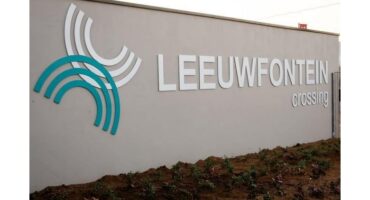 White and teal arch design next to "LEEUWFONTEIN crossing" on a beige wall, with small plants at the base. Emphasis on modernity and calmness.
