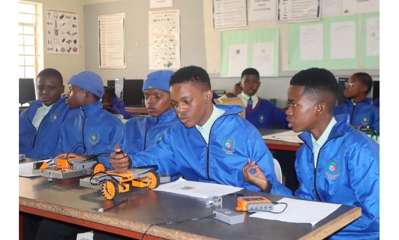 Students in blue jackets work intently on small robotic vehicles in a classroom, collaborating and focused, with educational posters on the walls.