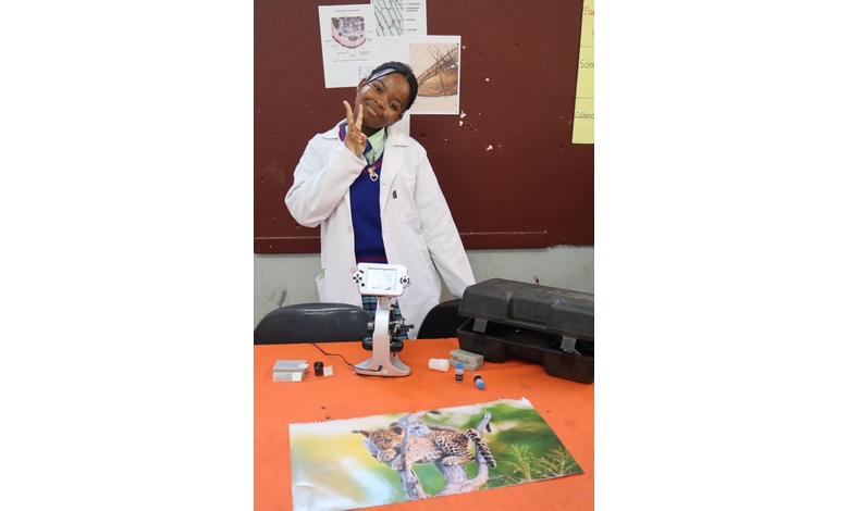 A person in a lab coat smiles and gestures peace signs beside a microscope on an orange table. Charts and a cheetah photo are in the background.